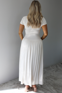 Woman wearing a light sand colored maxi length dress that has a fitted bodice with lace detailing in the center of the bust, a drop waist design, short sleeves, and a flowy skirt. The photo is taken indoors against a white wall with white marble flooring. The photo shows the back of the dress. 