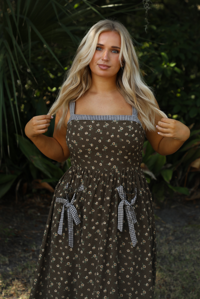 Woman model wearing a brown dress that has two different patterns. It has a multi-colored floral pattern throughout a brown and white checkered pattern at the neckline, straps, and bow details at the pockets. The dress has a fitted bodice, with a cinched waistline, functional pockets in the front, and a long length skirt that stops just above the ankle on the model. The photo shows the bodice, straps, and upper skirt in more detail. The photo is taken outdoors with greenery in the background.