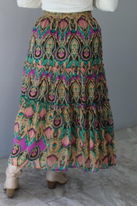 A woman, whose face is not showing, is shown from behind, wearing a long sleeved cream top and multi-colored maxi length skirt paired with knee high sand colored ankle boots, standing indoors against a white wall with white marble flooring. 