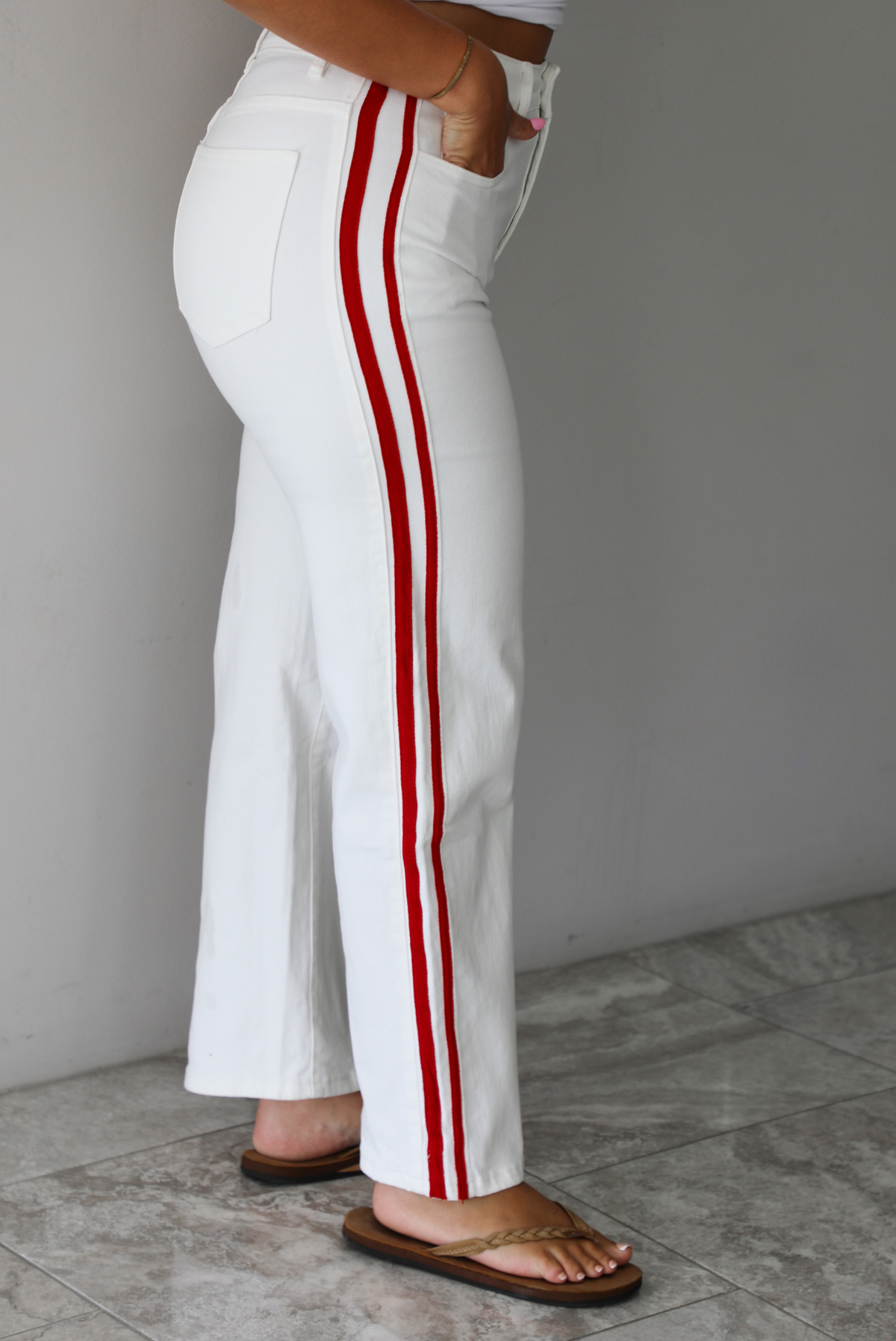 Woman wearing a white tank with white denim jeans with red stripes on either side  indoors against a white wall with white marble flooring. The photo shows a closeup of the side of the pants. 