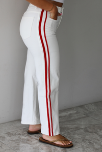 Woman wearing a white tank with white denim jeans with red stripes on either side  indoors against a white wall with white marble flooring. The photo shows a closeup of the side of the pants. 