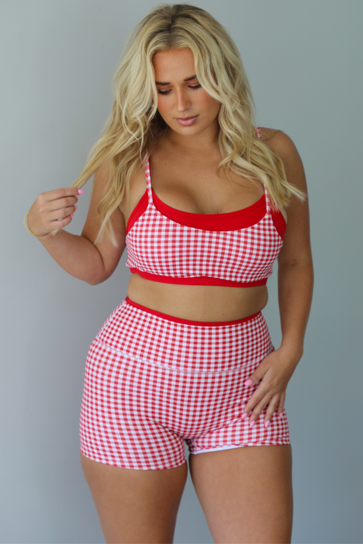 A woman modeling a red and white gingham two-piece athletic set featuring a sports bra and high waisted biker shorts with solid red trim indoors against a white wall.