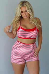 A woman modeling a red and white gingham two-piece athletic set featuring a sports bra and high waisted biker shorts with solid red trim indoors against a white wall.