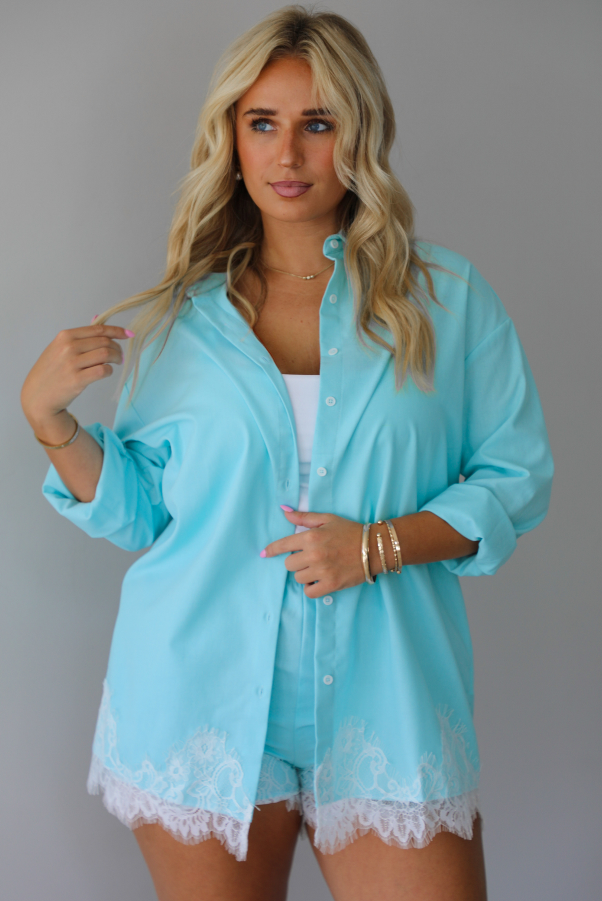 A woman stands indoors against a white wall wearing a light aqua blue matching lounge set consisting of a long open-front button-down and shorts with white lace trim, over a white tank top.