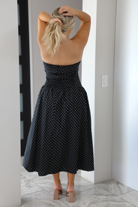 Back of a woman model wearing a strapless/sleeveless, black with white polka dots, maxi length dress indoors. It has a fitted bodice with a flowy skirt. 