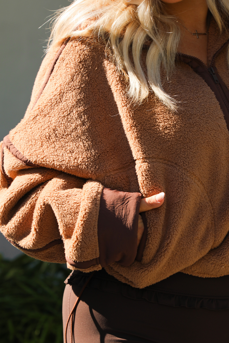 Woman model wearing a tan and brown quarter zip sweater with dark brown athleisure pants. The photo is taken outdoors against some greenery and a white wall. The photo showcases the sweater close up and the pocket on the side. 