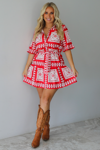 A woman modeling a red and white patterned short-sleeve mini dress with a tie waist, paired with brown cowboy boots, standing indoors against a white wall with white marble flooring. 