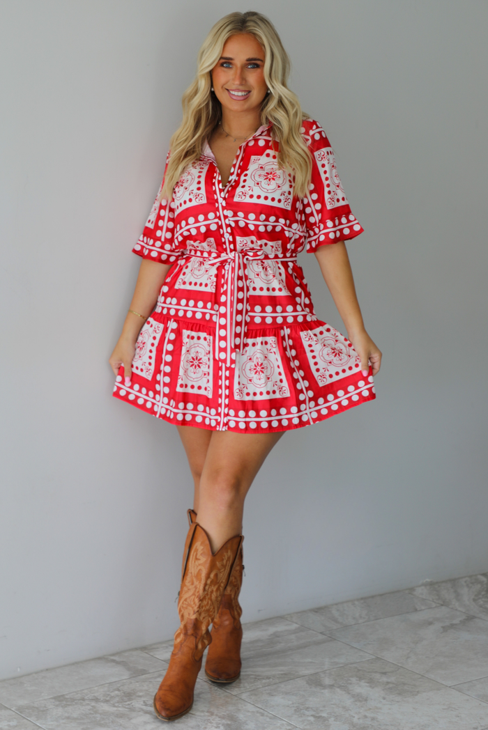 A woman modeling a red and white patterned short-sleeve mini dress with a tie waist, paired with brown cowboy boots, standing indoors against a white wall with white marble flooring. 