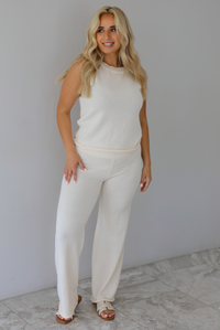 Woman wearing a cream two piece set featuring a high-neck sleeveless top and matching mid-rise pants indoors against a white wall with white marble flooring. 