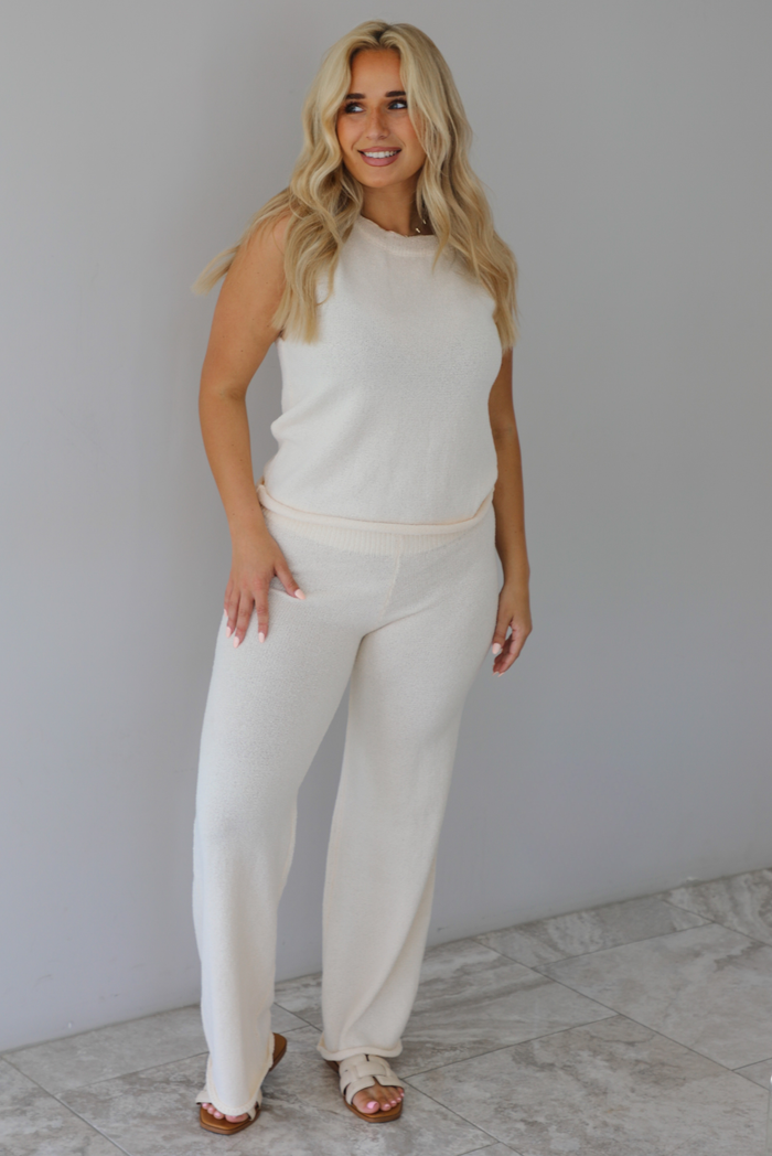 Woman wearing a cream two piece set featuring a high-neck sleeveless top and matching mid-rise pants indoors against a white wall with white marble flooring. 