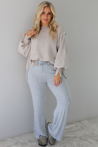 Woman wearing sand colored oversized sweatshirt with exposed seam details and a high-low design with heather grey casual pants. The outfit is styled with grey slip on shoes. The photo is taken indoors against a white wall with white marble flooring. 