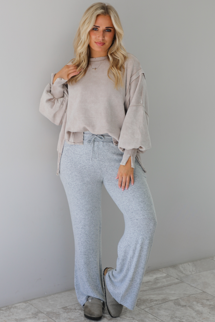 Woman wearing sand colored oversized sweatshirt with exposed seam details and a high-low design with heather grey casual pants. The outfit is styled with grey slip on shoes. The photo is taken indoors against a white wall with white marble flooring. 