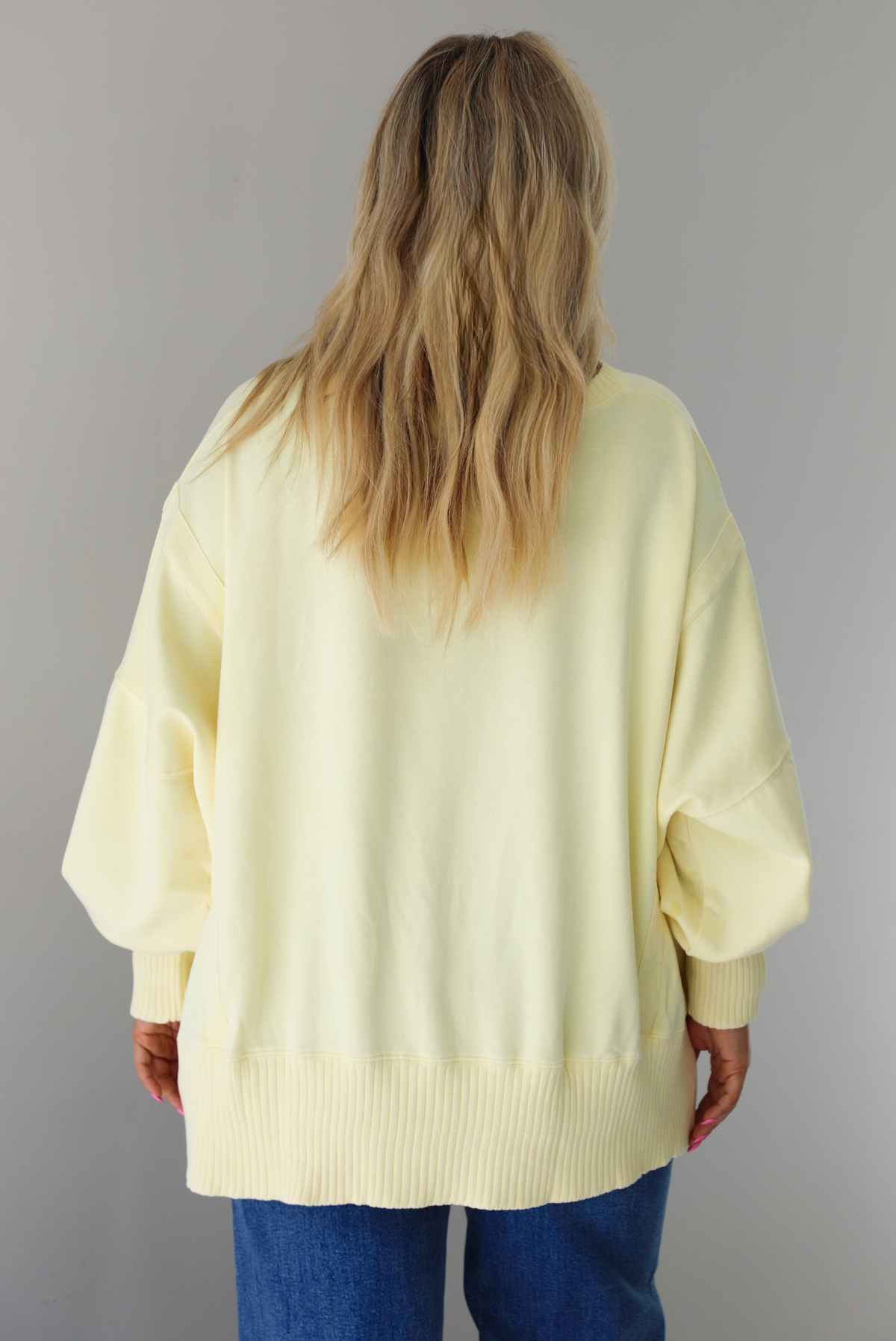 Woman wearing a pale yellow oversized sweater that's untucked and paired with dark wash denim. The photo shows the full length of the sweater.  The photo is taken indoors against a white wall.