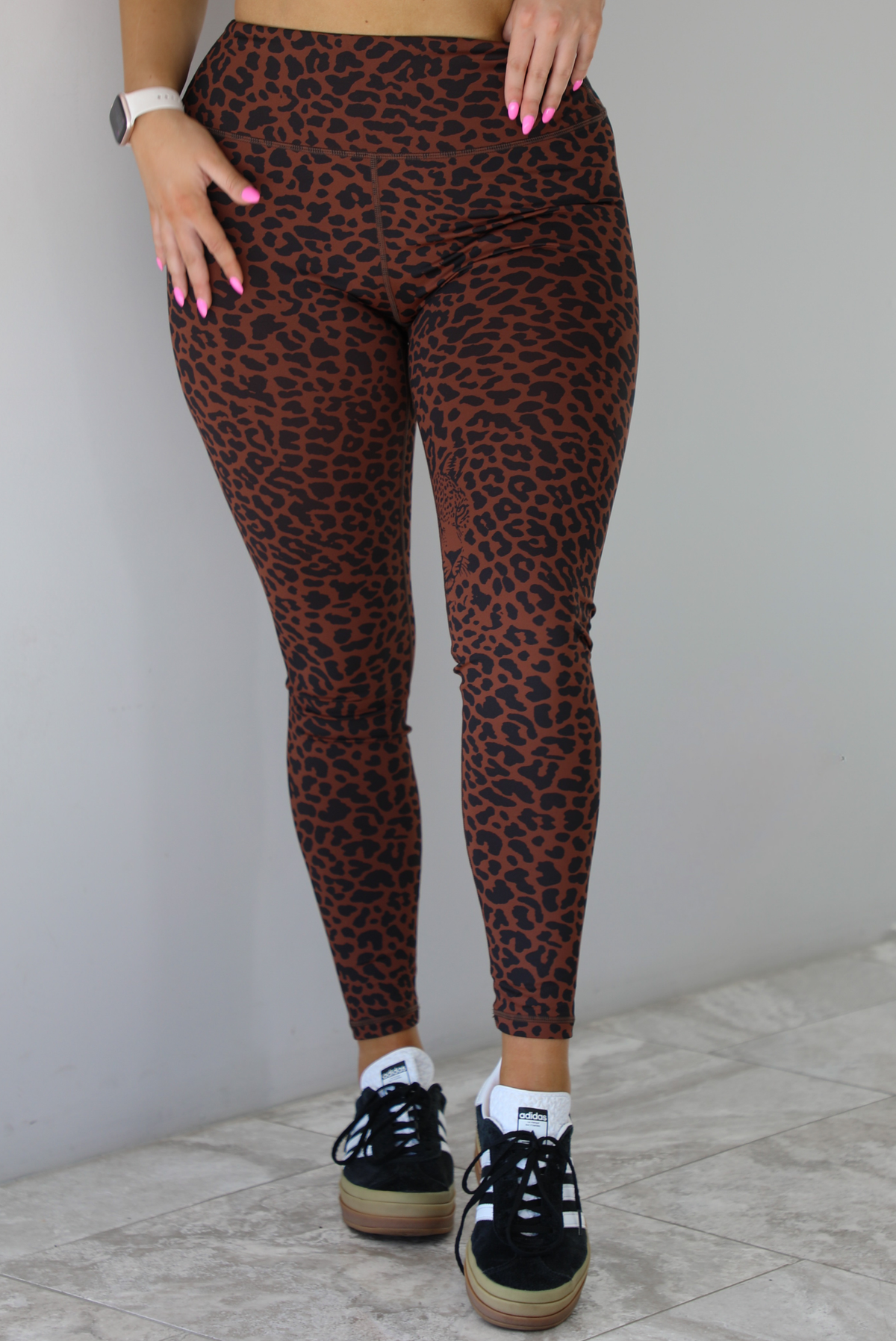 Woman wearing a brown and white leopard print two piece athleisure set that consists of a sports bra and high waisted leggings. The photo shows a closeup of the leggings. The photo is taken indoors against a white wall with white marble flooring. 