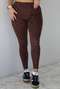 Woman wearing a brown and white leopard print two piece athleisure set that consists of a sports bra and high waisted leggings. The photo shows a closeup of the leggings. The photo is taken indoors against a white wall with white marble flooring. 