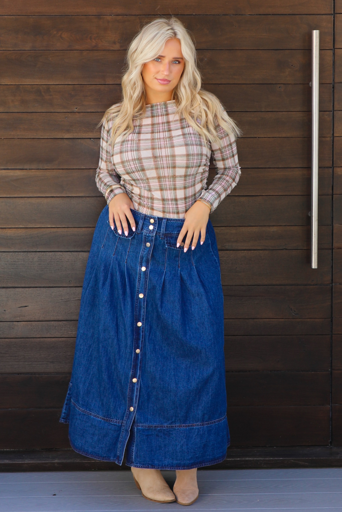 A woman wearing a beige and brown plaid long-sleeve top and a high-waisted blue denim button-front midi skirt, standing outdoors against a dark wooden door. 