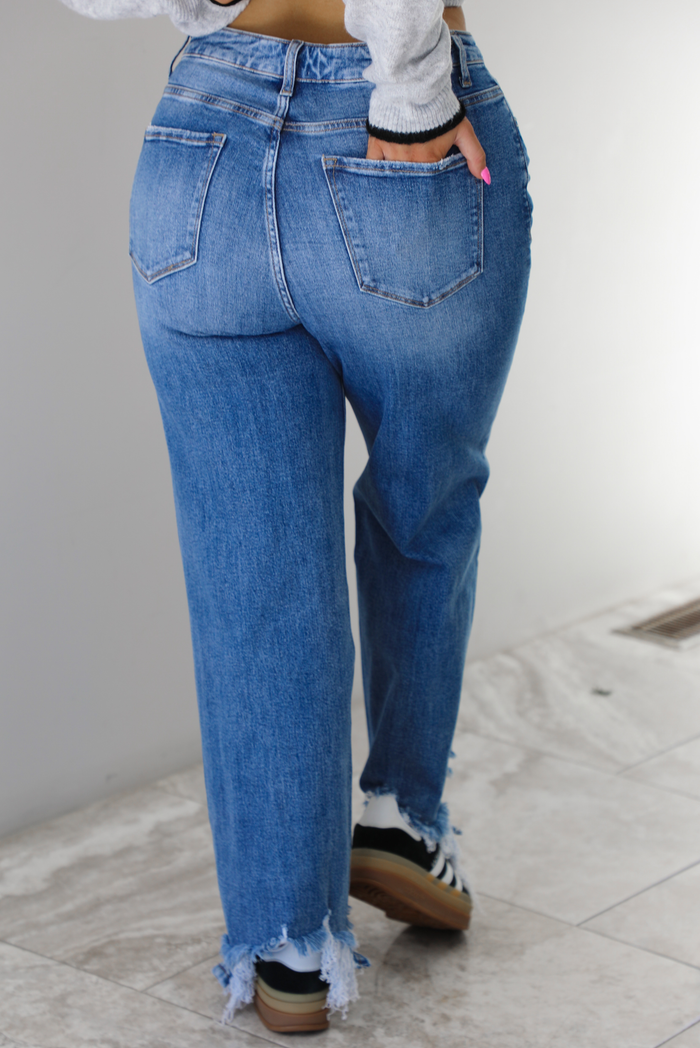 Woman wearing a medium wash denim that are high waisted, have functional front and back pockets, functional button and zipper closures, and have a raw, distressed hem. The photo shows the back of jeans, and the models hand is in the back right pocket. The photo is taken indoors against a white wall with white marble flooring.