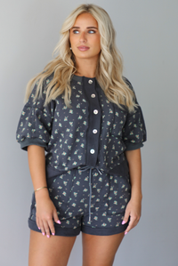 Woman wearing a charcoal with a multi-colored floral two piece set featuring an oversized top with puffed sleeves and shorts indoors against a white wall. The photo shows a close up of the set, showing the functional button closures on the front of the top and the elastic and drawstring waistline on the shorts. 