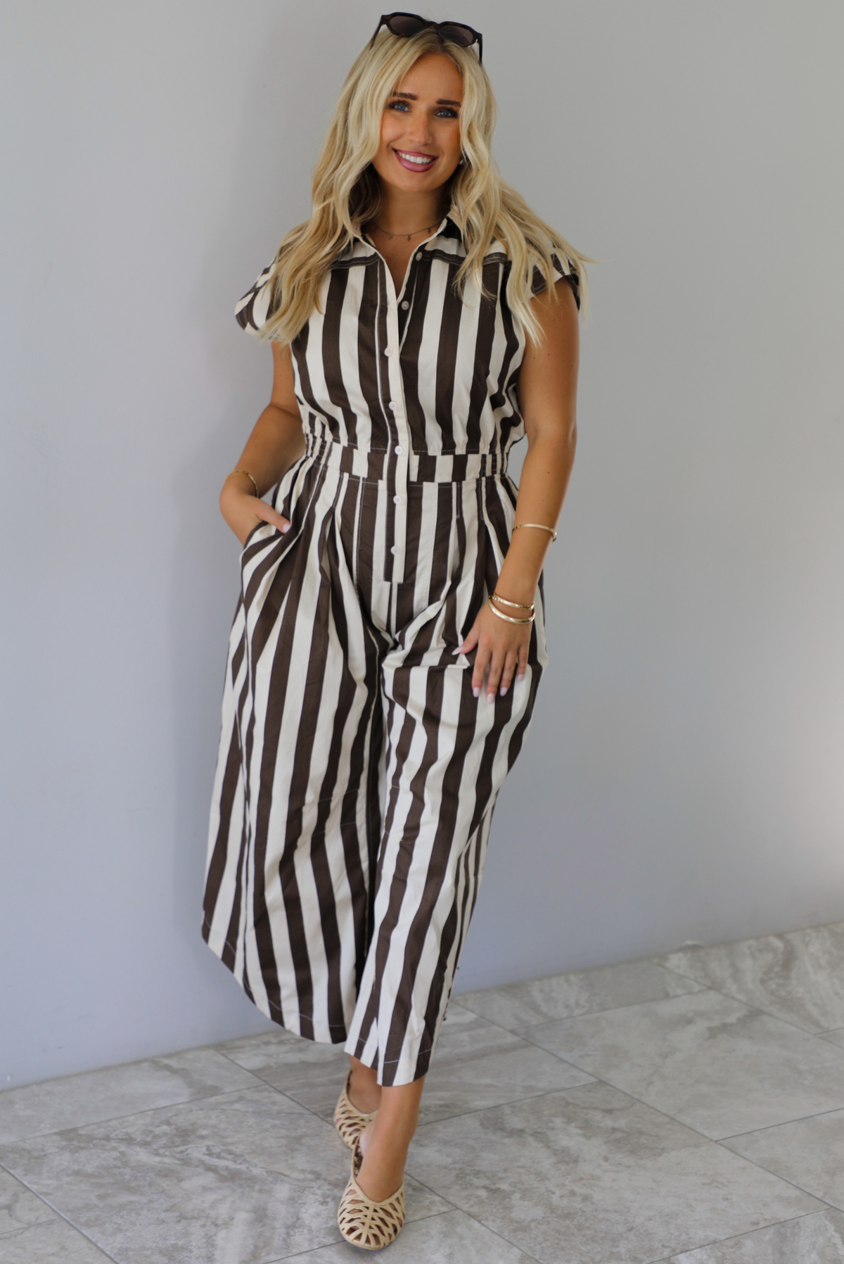 A woman wearing a brown and white striped patterned jumpsuit with a collared neckline, functional button closures in the front, a cinched waistline, and barrel-style bottoms, standing indoors against a white wall with white marble flooring. 