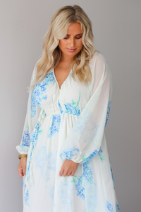 Closeup of a woman modeling a white maxi dress with a blue floral print and a sheer, flowing overlay, focusing on the bodice of the dress with its cinched waistline, standing indoors against a white wall. 