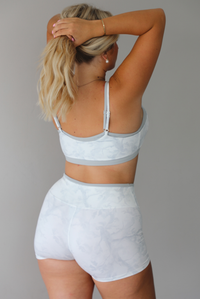 A woman shown from behind wearing a matching grey and white camo activewear set consisting of a sports bra and high-waisted shorts, standing indoors against a white wall.