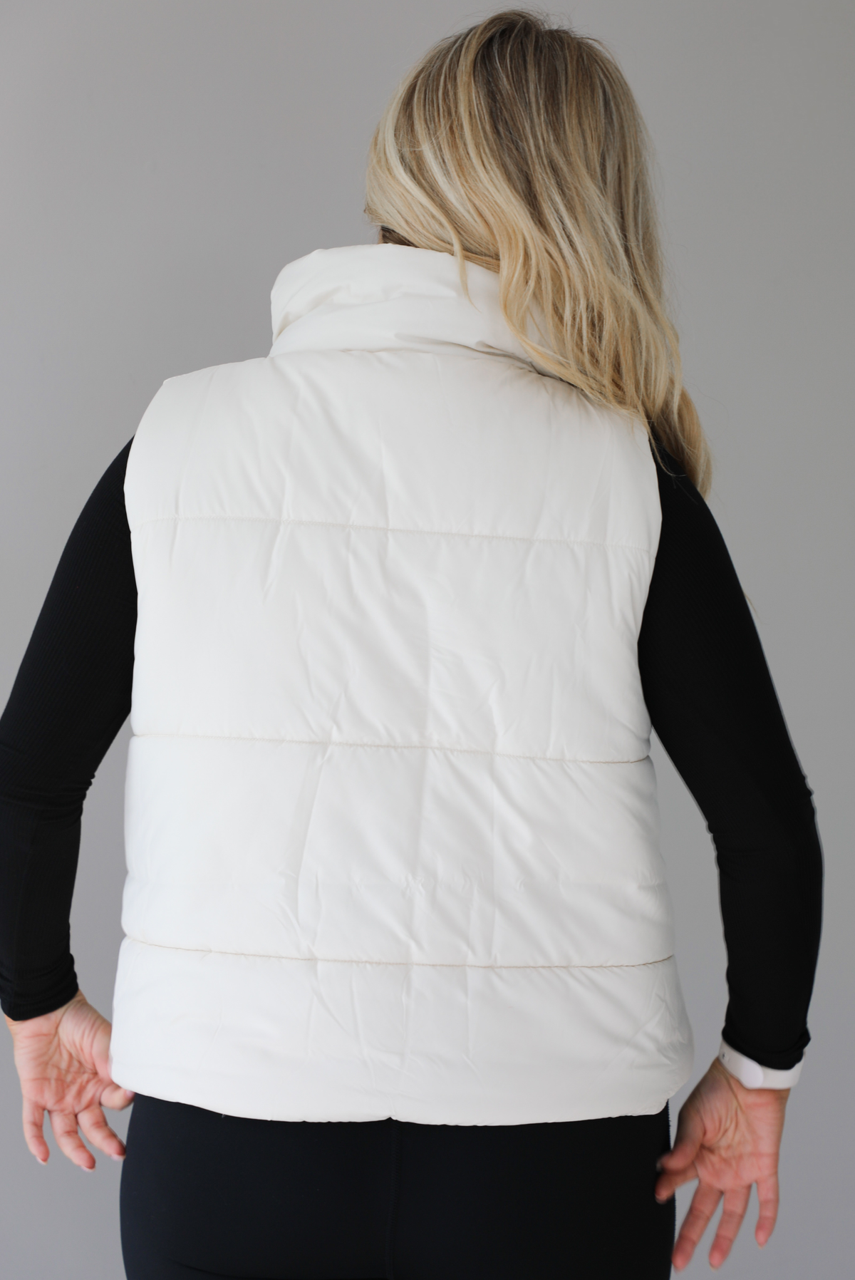 Woman wearing a light sand colored puffer vest with a fitted black long sleeved top and fitted leggings. The vest has functional button closures at the top, a zipper closure in the front, and functional pockets on either side. The photo shows the back of the vest. The photo is taken indoors against a white wall with white marble flooring. 
