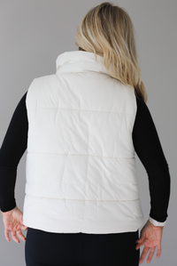 Woman wearing a light sand colored puffer vest with a fitted black long sleeved top and fitted leggings. The vest has functional button closures at the top, a zipper closure in the front, and functional pockets on either side. The photo shows the back of the vest. The photo is taken indoors against a white wall with white marble flooring. 