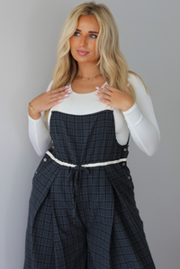 A woman modeling a white long-sleeve top and dark gray plaid wide-leg overalls, standing indoors against a white wall.