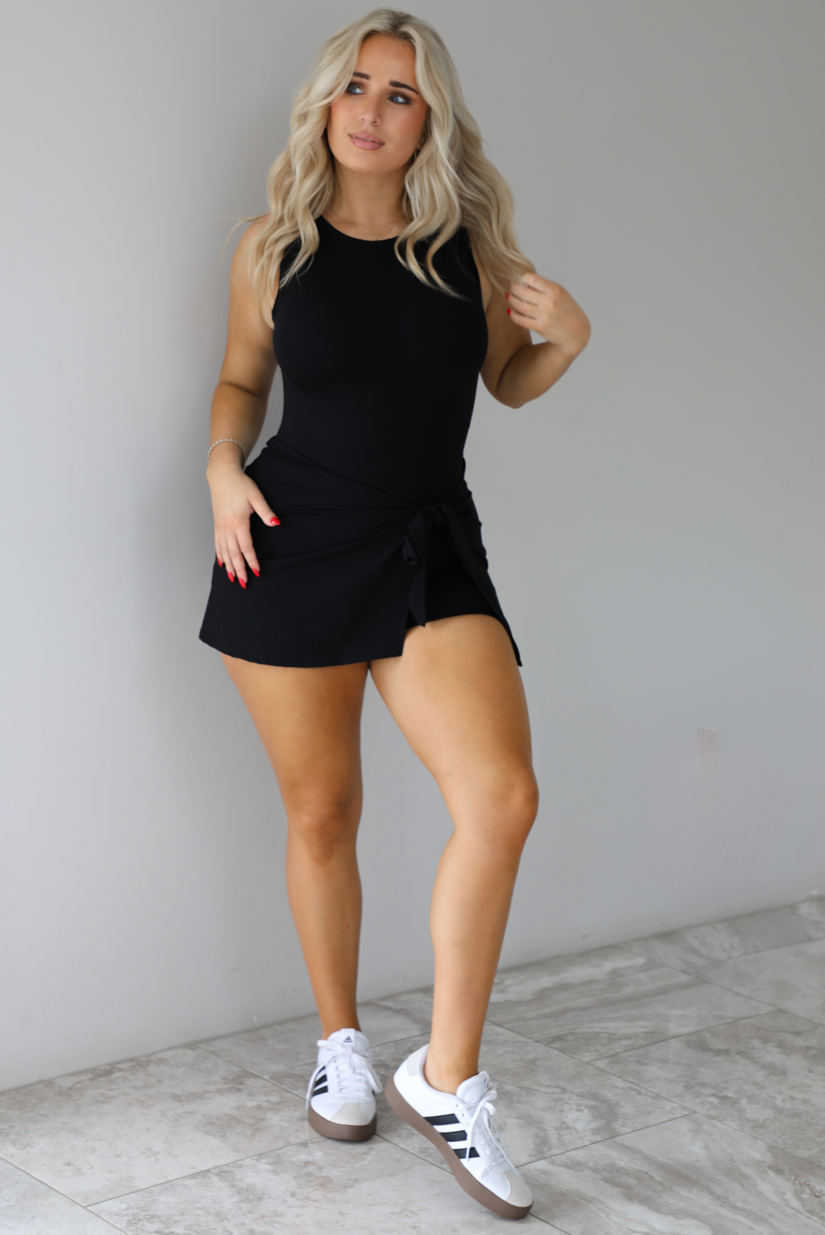 Woman wearing a black athleisure romper that has a wrap-style skirt detail that wraps around and ties in the front on the left side. It's styled with white sneakers that have black stripes down the side. The photo is taken indoors against a white wall with white marbled flooring. 