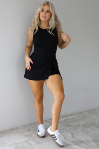 Woman wearing a black athleisure romper that has a wrap-style skirt detail that wraps around and ties in the front on the left side. It's styled with white sneakers that have black stripes down the side. The photo is taken indoors against a white wall with white marbled flooring. 