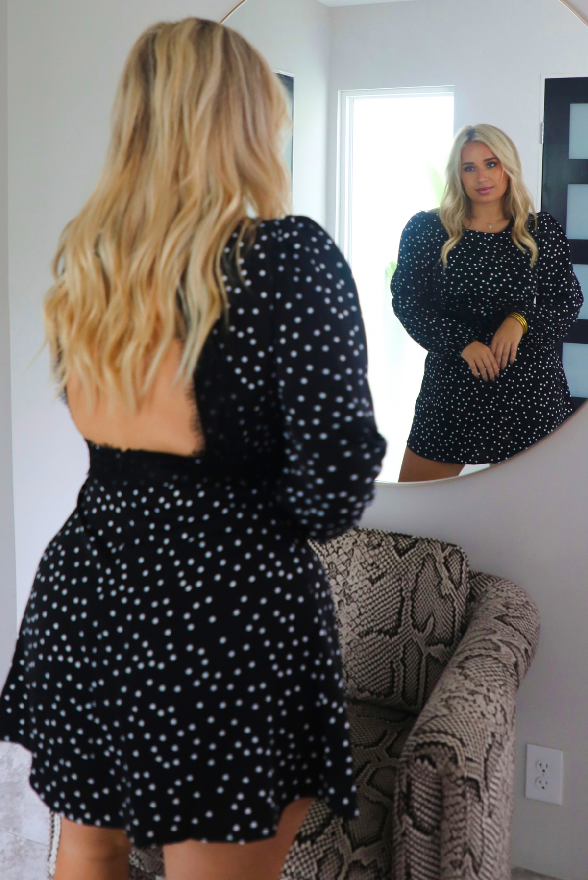 A woman wearing a black dress with white polka dots that has long sleeves, a high neckline, a cinched waistline, an open back, and a flowy skirt. The photo is taken behind the model as she looks into a mirror, showing both the front and back of the dress. The photo is taken indoors with a snake pint armchair in front of the model.