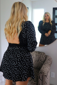 A woman wearing a black dress with white polka dots that has long sleeves, a high neckline, a cinched waistline, an open back, and a flowy skirt. The photo is taken behind the model as she looks into a mirror, showing both the front and back of the dress. The photo is taken indoors with a snake pint armchair in front of the model.