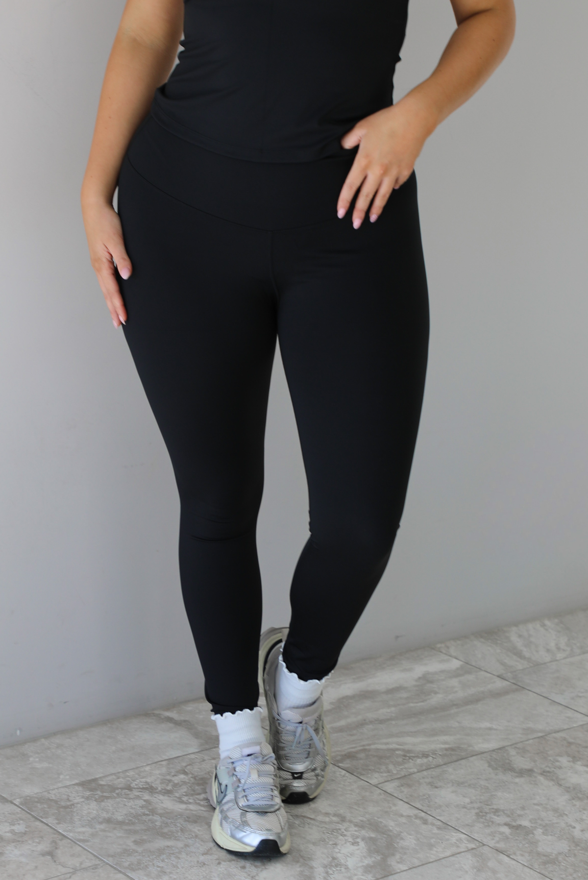 Woman wearing a fitted two piece black set featuring a short sleeved top and fitted leggings. The photo shows a closeup of the pants, which is paired with white ankle socks and silver sneakers.  The photo is taken indoors against a white wall and white marble flooring. 