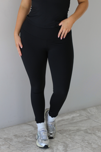 Woman wearing a fitted two piece black set featuring a short sleeved top and fitted leggings. The photo shows a closeup of the pants, which is paired with white ankle socks and silver sneakers.  The photo is taken indoors against a white wall and white marble flooring. 
