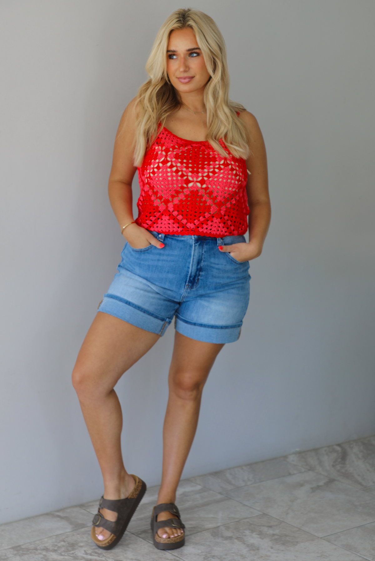 Woman wearing a pink and red loose knit crochet cropped top with light wash denim shorts indoors against a white wall with white marble flooring. 