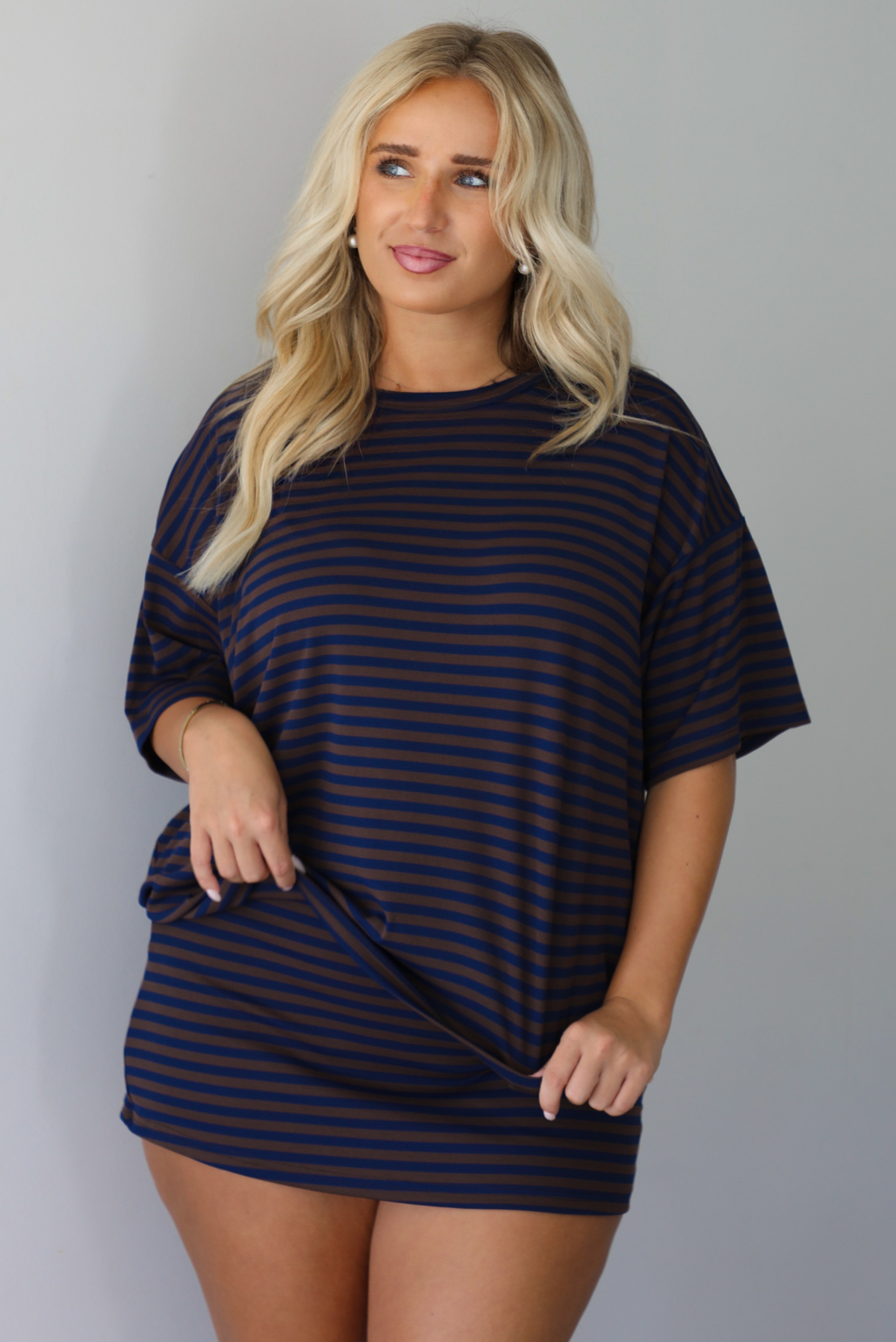 A woman wearing a navy and brown striped set featuring an oversized top and mini skirt, standing indoors against a white wall.