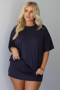 A woman wearing a navy and brown striped set featuring an oversized top and mini skirt, standing indoors against a white wall.
