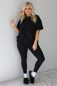 Woman wearing a black two piece set featuring an oversized t-shirt and fitted leggings. The set is paired with high white ankle socks and black slip on shoes. The photo is taken indoors against a white wall with white marble flooring. 