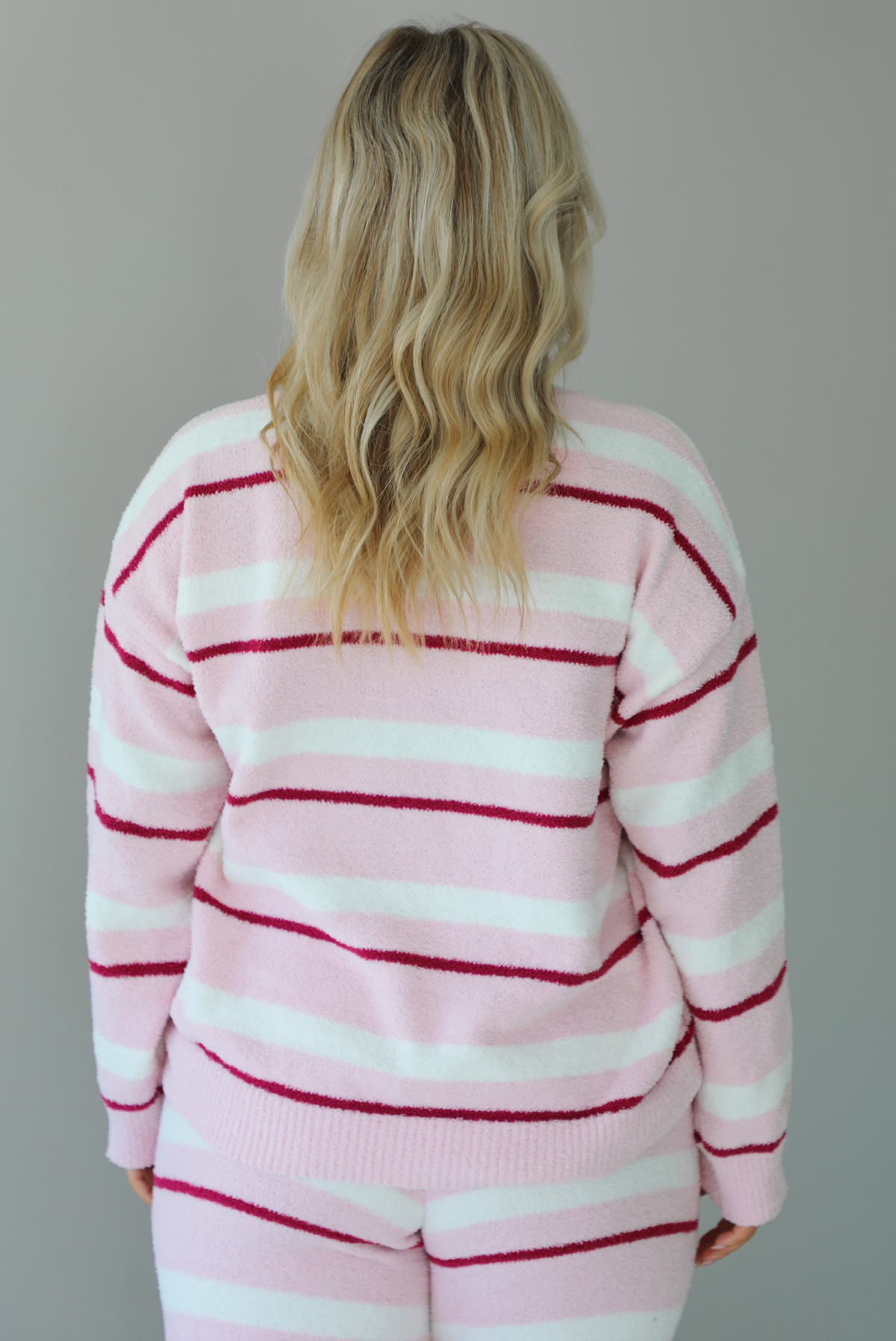 Woman wearing a pink set with a darker pink and white striped design. The set consists of an oversized sweater top and high waisted pants. The photo shows a closeup of the back of the sweater, untucked. The photo is taken indoors against a white wall. 