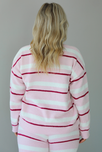 Woman wearing a pink set with a darker pink and white striped design. The set consists of an oversized sweater top and high waisted pants. The photo shows a closeup of the back of the sweater, untucked. The photo is taken indoors against a white wall. 