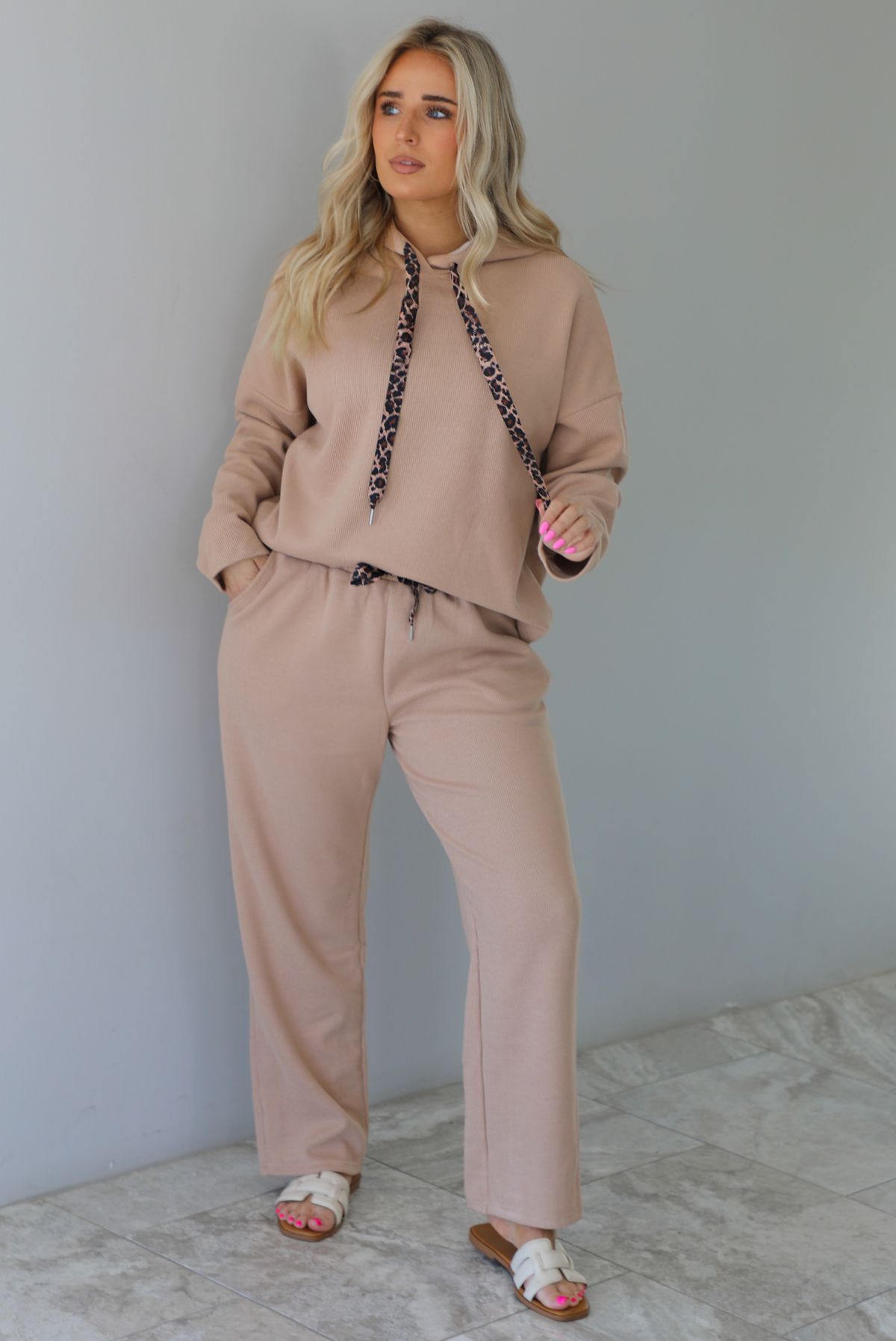 Woman wearing a tan two piece set with a hoodie top and sweatpants. The photo is taken indoors against a white wall with white marble flooring. 
