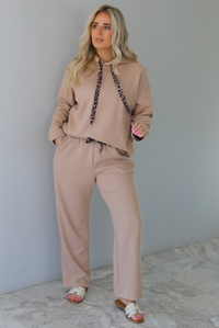 Woman wearing a tan two piece set with a hoodie top and sweatpants. The photo is taken indoors against a white wall with white marble flooring. 