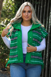 Woman model wearing a green, black, and white striped vest that has a sherpa texture to it, functional button closures in the front, and tie closures on either side, It is paired with a white long sleeved top, a medium wash pair of denim jeans, and tan ugg boots. The photo is taken outdoors with a metal gate and greenery in the back. The vest is shown close up, displaying the buttons unclasped.