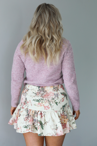 Woman wearing a mauve colored ribbed cardigan that has gold button closures along the front with a cream and multi-colored floral print patterned skirt. The photo is taken indoors against a white wall. The photo shows the back of the cardigan and skirt. 