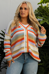 Woman model wearing an orange, lavender, yellow, muted green, blue, and white striped oversized cardigan with functional button closures in the front and an oversized fit with a light wash flared hem denim. The photo is taken outdoors against a white wall and greenery in the back. The photo shows a close up of the cardigan that's styled tucked in the front.