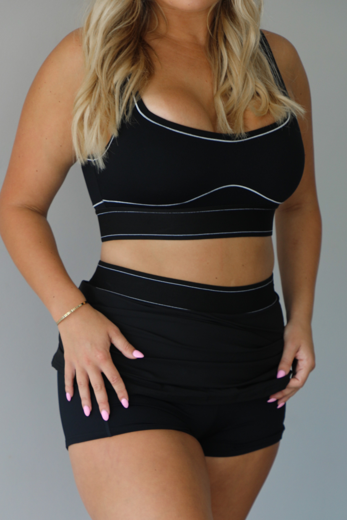 A blonde woman modeling a black two-piece activewear set consisting of a scoop-neck sports bra and a high-waisted mini skirt, both featuring thin white contrast piping details, indoors against a white wall. The model is holding up the skirt, revealing the shorts liner underneath. 