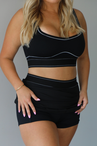 A blonde woman modeling a black two-piece activewear set consisting of a scoop-neck sports bra and a high-waisted mini skirt, both featuring thin white contrast piping details, indoors against a white wall. The model is holding up the skirt, revealing the shorts liner underneath. 
