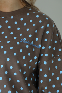 Woman wearing a brown sweater that has a blue polka dot print pattern throughout with blue embroidered writing on the left side that reads 'Trust The Process.' The photo shows a closeup of the letting on the left side. The photo is taken indoors against a white wall.