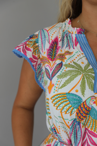 A woman wearing a short sleeved midi dress with a colorful tropical leaf print and light blue trim indoors against a white wall. The photo shows a closeup of the blue scalloped trim detailing on the sleeves. 