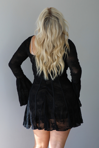 A woman shown from behind wearing a black, long sleeved mini dress with a low neckline and a slightly flowy skirt, with a zipper closure in the back, standing indoors against a white wall.